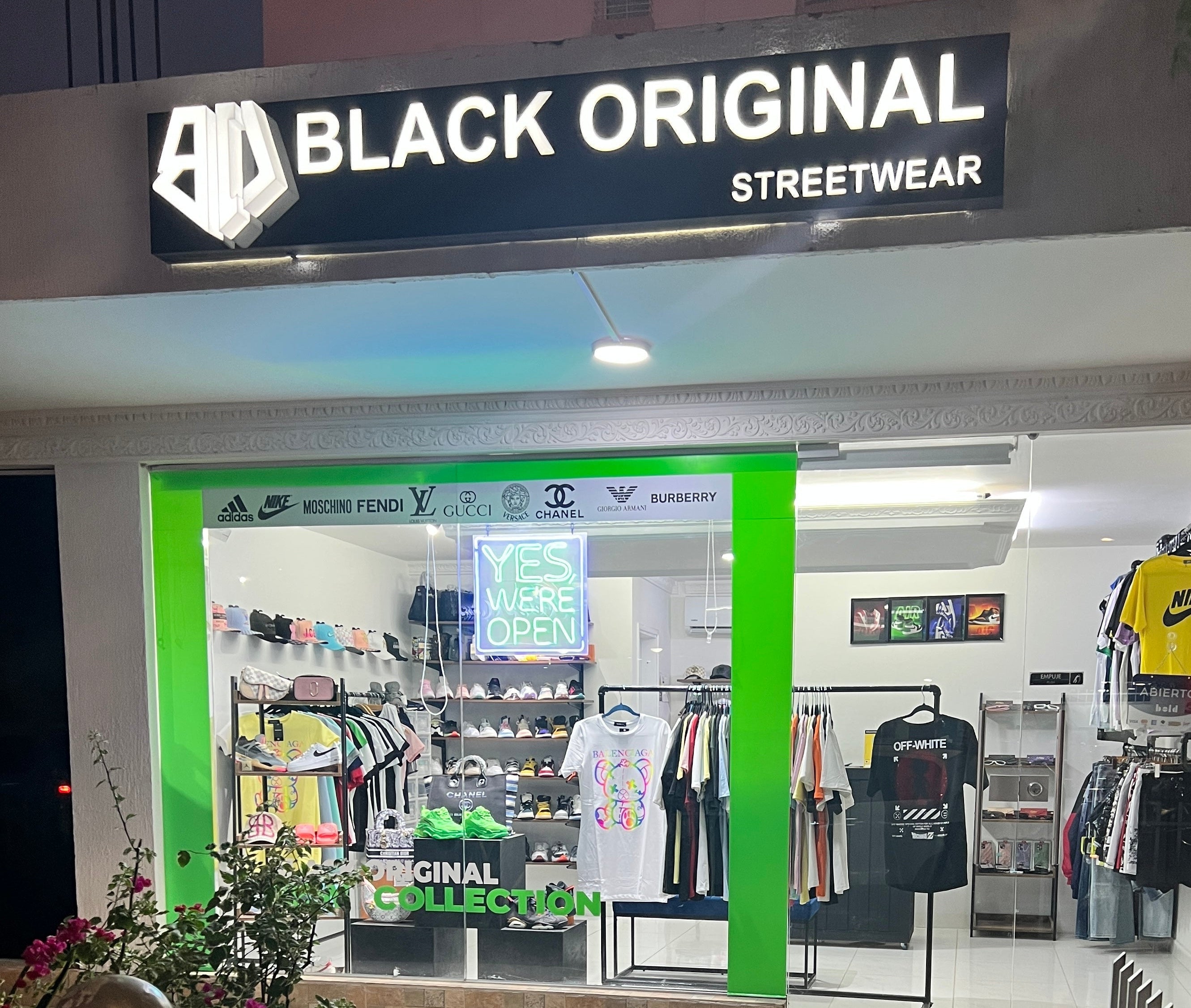 Black Original Streetwear storefront at night with a large illuminated black sign above a bright green-framed window display. Inside, clothing racks, shelves, and a neon sign reading 'YES WE'RE OPEN' are visible, with a yellow Nike shirt hanging on the right.